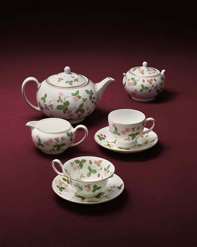 A selection of five Wild Strawberry teaware pieces presented on a red studio background. The selection features a teapot, cream jug, two teacups and saucers, and a sugar bowl.