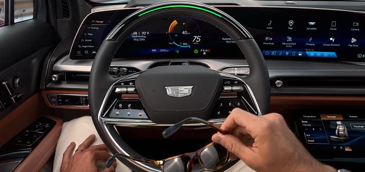 Front passenger seat view of the 2026 Cadillac VISTIQ’s steering wheel with Super Cruise activated; the driver is relaxed, looking ahead, and holding their sunglasses.