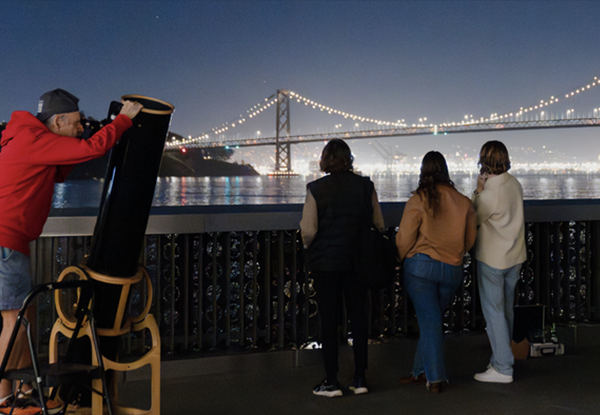 People looking at the Bay Bridge at night, and a man on the side looking through a telescope.
