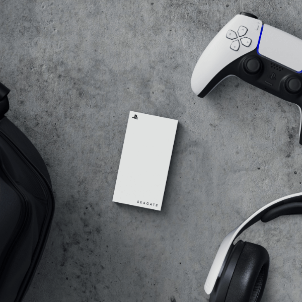 A white Seagate external hard drive is on a gray textured surface, next to a PlayStation controller, headphones, and a bag.