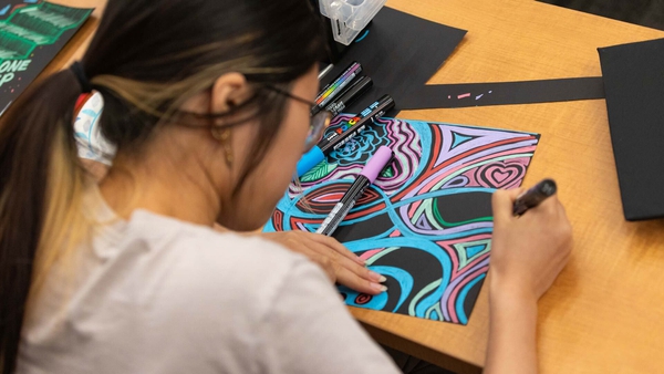 A student shown from behind bends over a pink, purple and black artwork she is coloring.