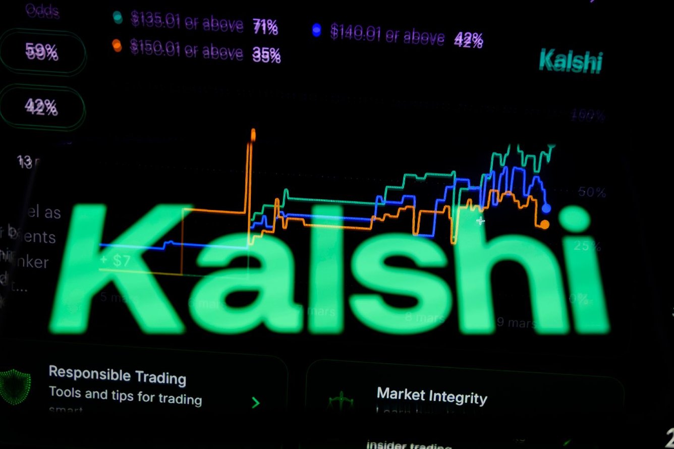 Photo of Kalshi's logo is displayed on a smartphone placed on a reflective surface onto which a betting curve is projected.