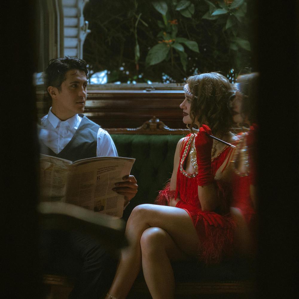 A man and woman in 1920's party garb sit on an upholstered bench together.