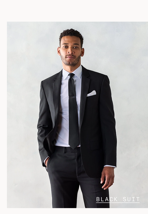 Man with short curly hair looking directly at camera with a neutral expression, wearing a black suit jacket over a white dress shirt with a black skinny tie and white pocket square, hand in pocket. BLACK SUIT