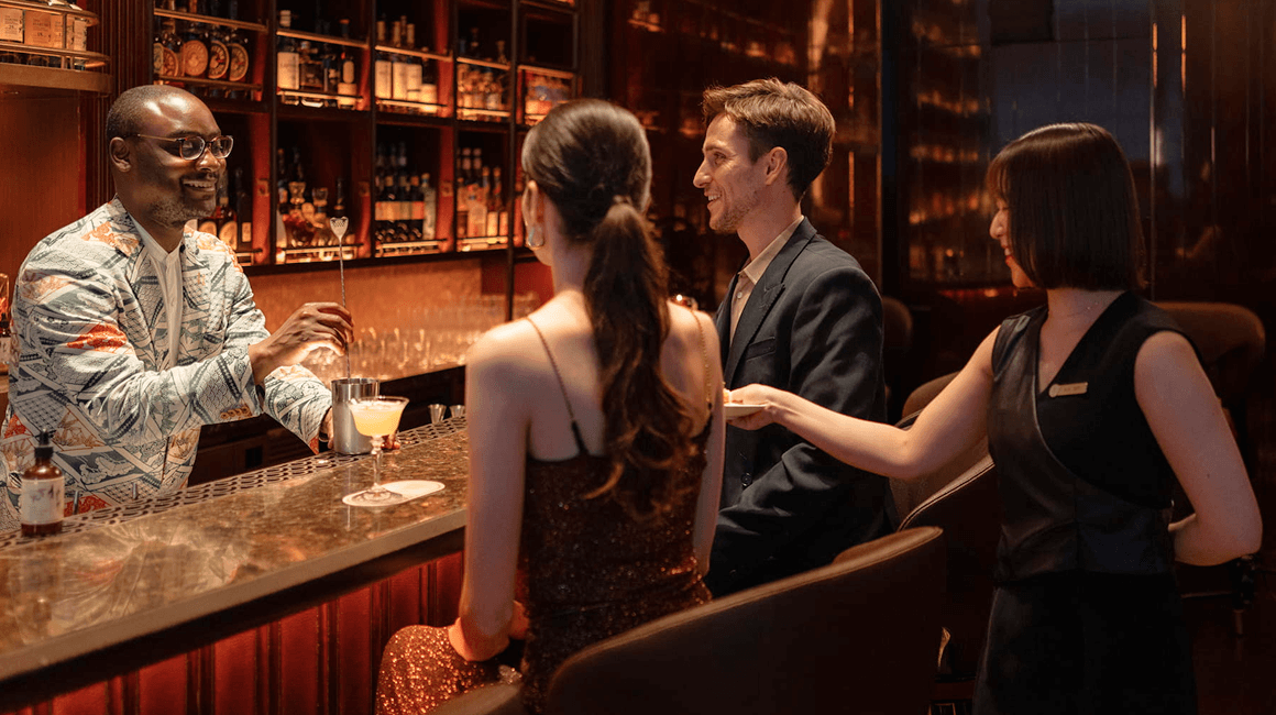 A bartender serves drinks to three well-dressed people at a dimly lit bar.