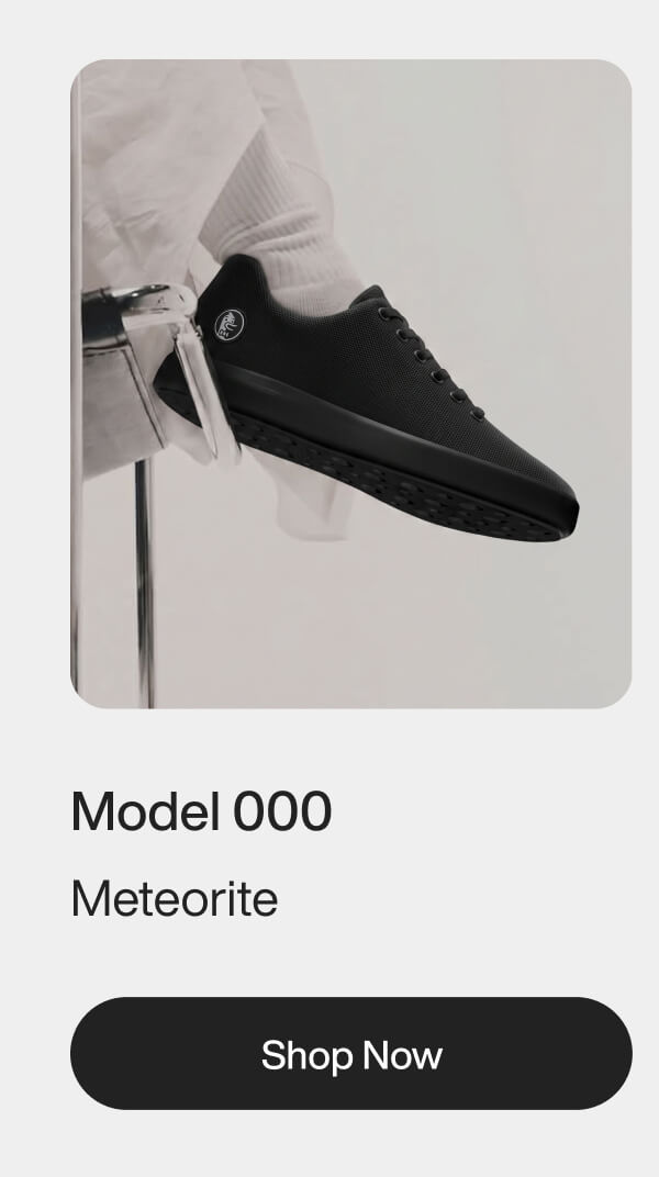 Black sneaker with a sleek design, labeled "Model 000 Meteorite," is worn by a person in white pants, perched on a stool. Modern and stylish tone.