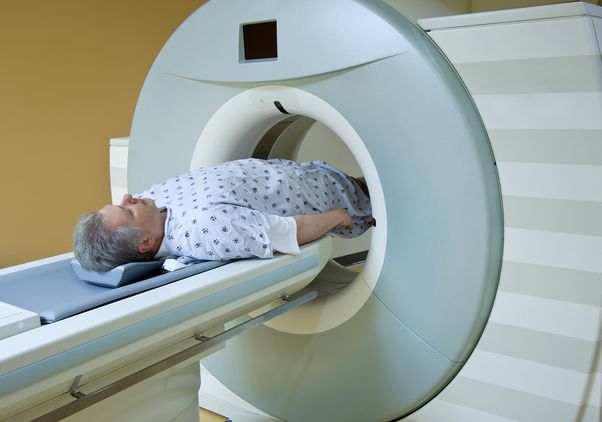A man with gray hair and a beard lies on his back on a scanner table, wearing a patterned hospital gown, as he is moved into the circular opening of a large MRI or CT scanner in a medical exam room.