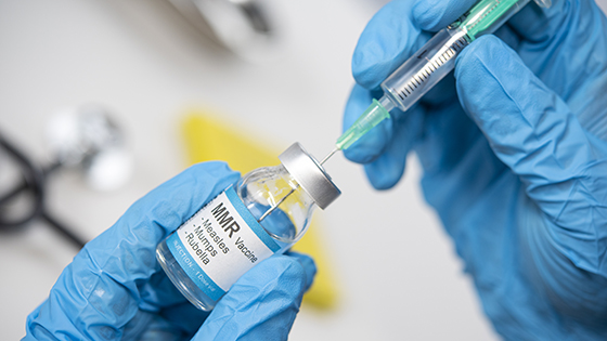 A person wearing blue latex gloves inserts a needle in a vile marked MMR. 