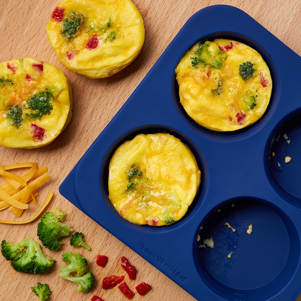 Egg bites made with broccoli, red bell pepper, and cheddar cheese cooked in the microwave using the Anyday Anytime Freezer Trays (½ cup size) in Blueberry, shown with fresh ingredients on a wooden surface.