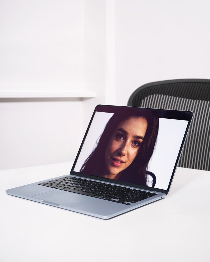 A laptop on a table, behind which is an empty office chair. On the laptop's screen is the smiling face of a woman with long dark hair and dark eyes.