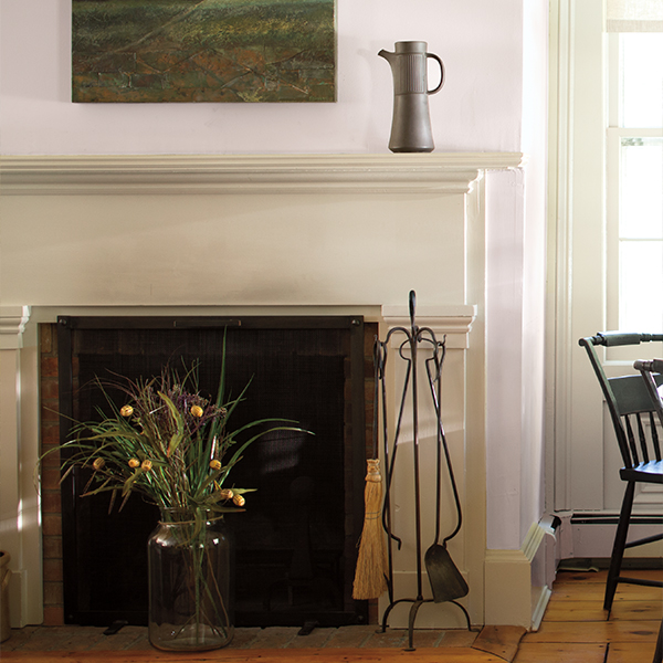 A living room with light lavender-painted walls, an off-white-painted fireplace and trim, with a rustic painting, and flowers in a vase.
