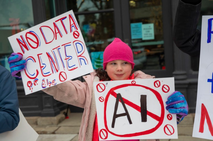 A child in a pink hat holds two handmade anti-data center signs at a protest.