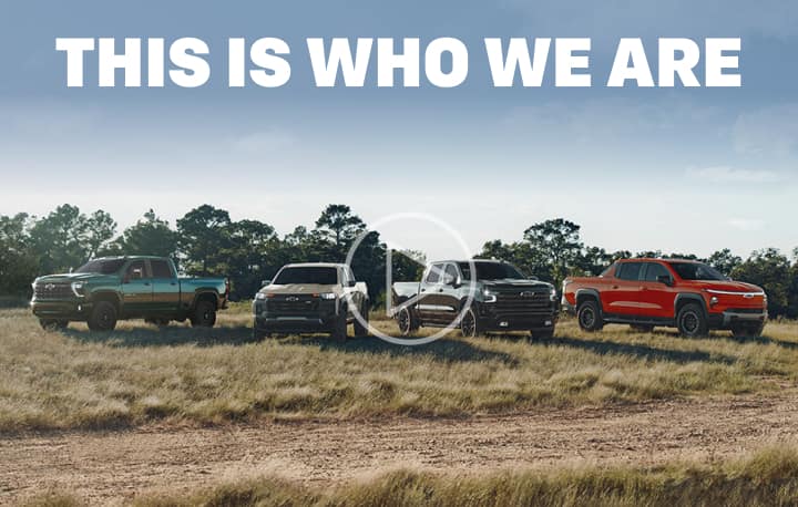This is who we are. A lineup of four Chevy trucks parked off-road in a field with trees in the background.