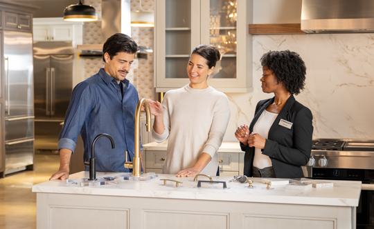 Three individuals standing in a kitchen, positioned near a sink and counter, engaged in conversation or activity.