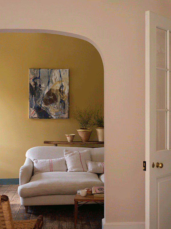 A GIF showing two scenes: first, a small cobblestone courtyard with green-painted brick walls, potted plants, and sculptural stone tables beside steps leading to a tall glass door; then a cosy living room with a mustard-yellow wall, light sofa with striped cushions, abstract artwork, and plants on a wooden console table.