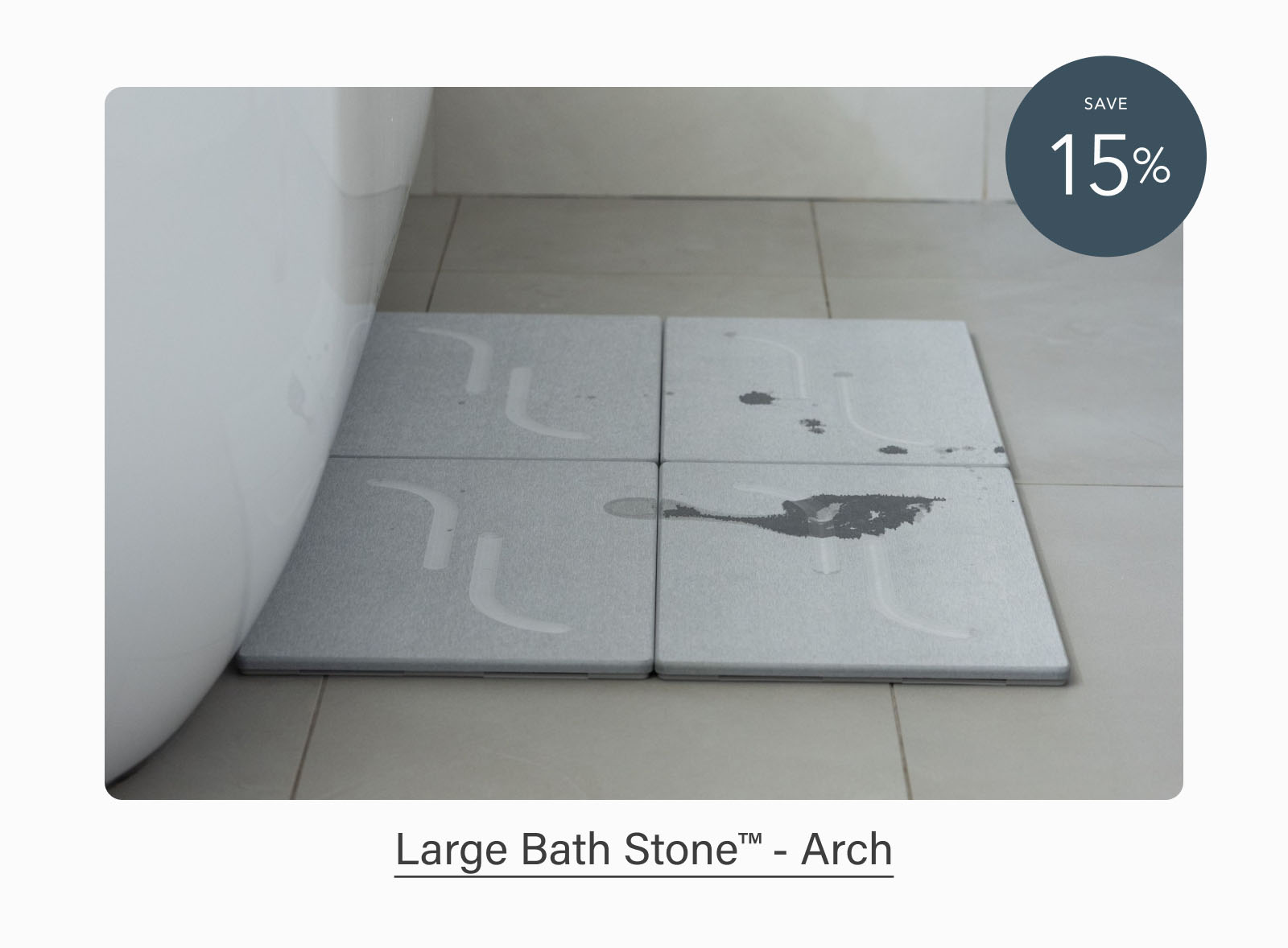 Light gray Dorai Large Bath Stone tiles arranged beside a bathtub, showing water evaporating from the surface. Badge indicates 15% savings. Text reads: "Large Bath Stone™ - Arch."