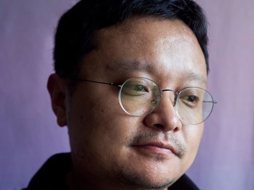 Close up of a figure who wears glasses, a dark shirt, and short hair. He looks slightly downwards away from the camera