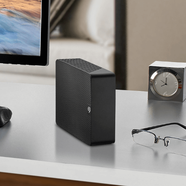 An expansion desktop hard drive sits on a tidy desk near a computer monitor, glasses, and a modern clock	