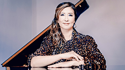 Rutgers professor Min Kwon is in seated pose, with her forearms resting on a black shine table surface with a raised piano lid in the background.