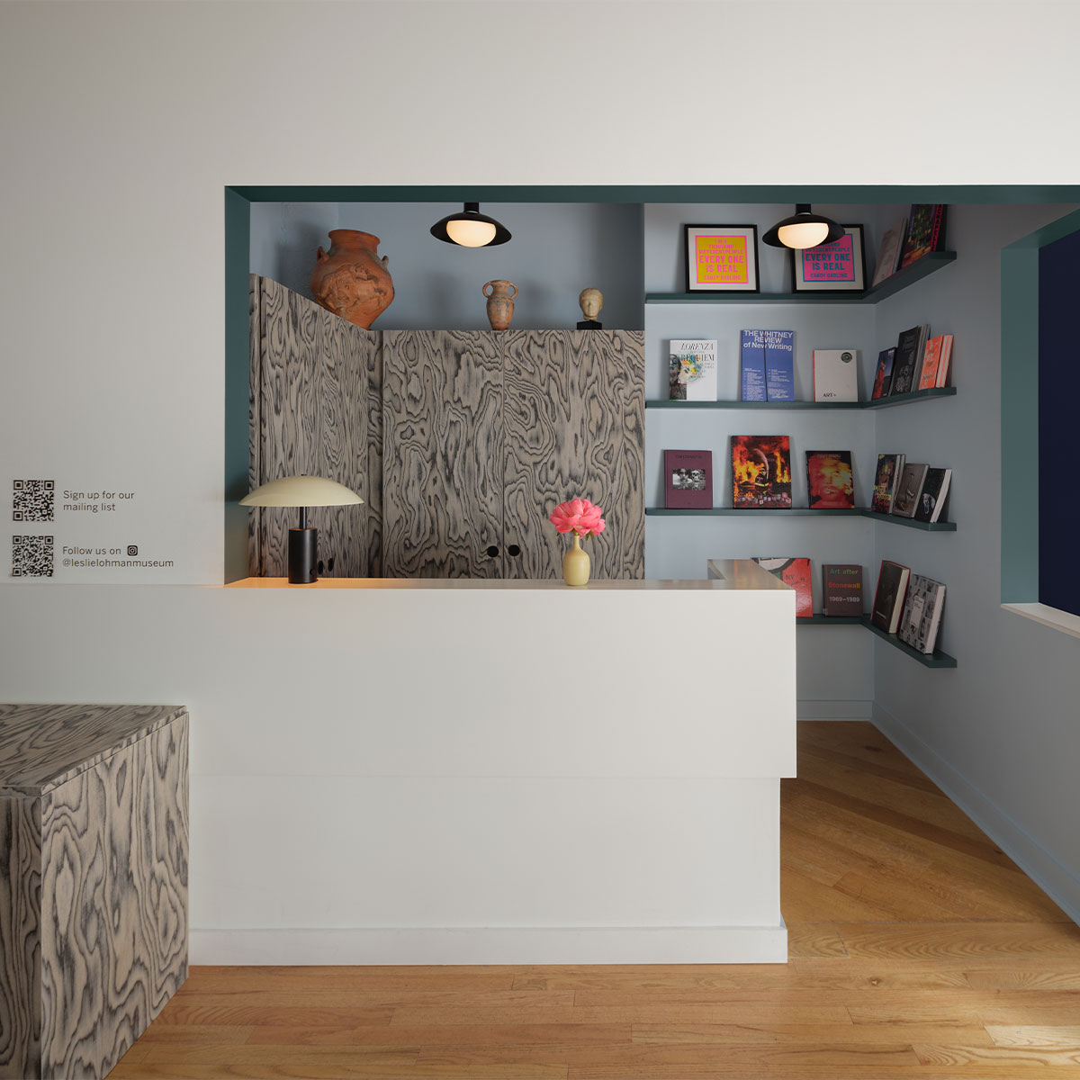 The reception area of the Leslie-Lohman Museum in New York. There’s a white desk built into the space with bookshelves on the wall in the recess behind it. The walls are painted in a light blue with a deeper blue accent on the edges. The colour combination creates a calm feel, like a cool sea breeze.