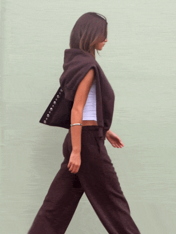Woman wearing a dark brown sleeveless hoodie and matching wide-leg pants with a white crop top underneath
