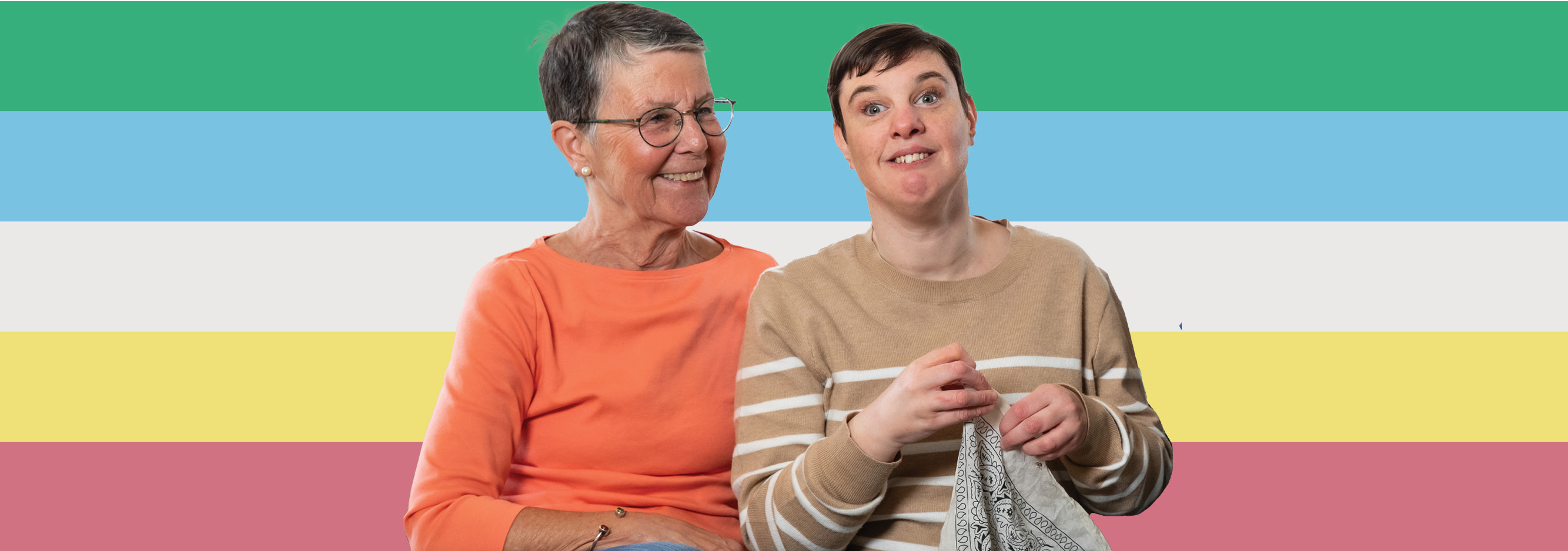 An older woman sits next to a younger woman with a disability, both smiling. There's a colorful striped background behind them, symbolizing disability pride.