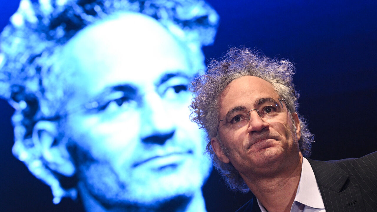 Palantir co-founder and CEO Alex Karp speaks at an event with his face projected on a screen in the background.