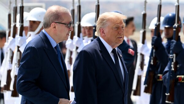 Turkish President Recep Tayyip Erdogan (left) and President Trump (right) stand next to each other. They are both wearing dark blue suits. Behind them are several people wearing uniforms and holding rifles upward. 