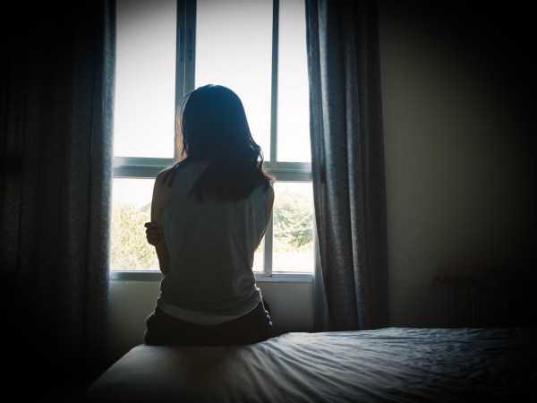 Sad person looking out window from darkened room on sunny day.