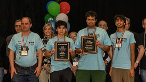 UMD computer science team stands on podium while wearing medals and holding up two plaques, with balloons behind them