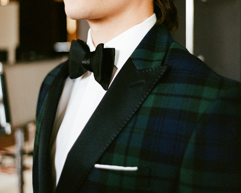 The image shows a person wearing a Black Watch tartan dinner jacket. This type of jacket is a stylish alternative to a traditional tuxedo jacket.  The Black Watch tartan is a well-known Scottish tartan, also known as the Government tartan.  It is characterized by its distinctive pattern of dark blue, green, and black stripes.  The jacket is often worn for formal events, such as New Year's Eve parties or important dinners.  The jacket in the image features a black bow tie and a white shirt, which are standard formal wear accessories. 