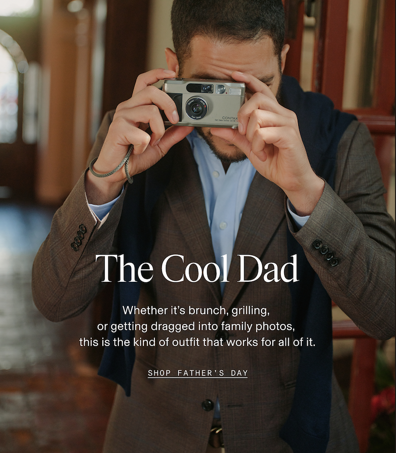 Man in a brown blazer and light blue button-down shirt holding a Contax film camera up to his face in a warmly lit interior setting. The Cool Dad. Whether it's brunch, grilling, or getting dragged into family photos, this is the kind of outfit that works for all of it. SHOP FATHER'S DAY