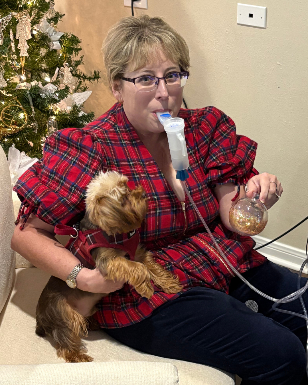 A fair-skinned woman holding her dog and a Christmas ornament while using her nebulizer