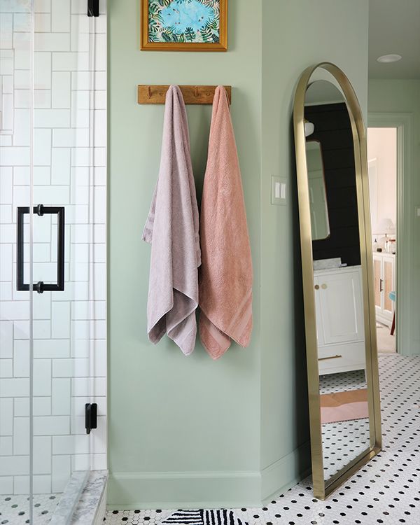 A mint green bathroom with a subway tile walk-in shower, a wooden hook with purple and pink towels, and a full-length, gold-rimmed mirror.