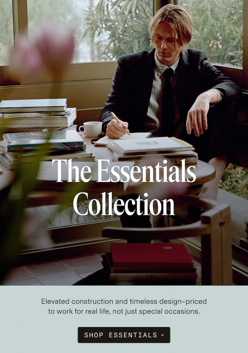 Man in a dark suit, striped shirt, and black tie writing at a desk surrounded by stacked books and a coffee cup, with greenery visible through a window behind him. Text reads: Elevated construction and timeless design – priced to work for real life, not just special occasions. SHOP ESSENTIALS →