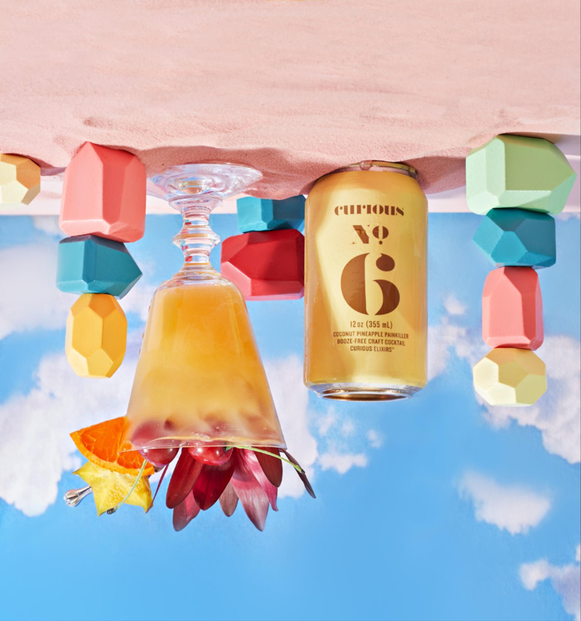 A can and garnished glass of Curious No. 6 alongside stacks of colorful blocks. All of these hang upside down off a pink, sandy surface into the blue sky. 