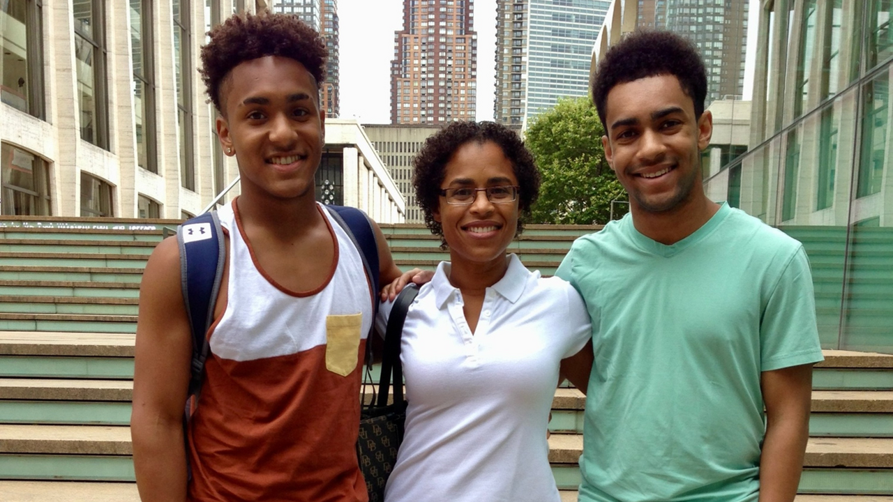A dark-skinned woman smiling and standing between her sons, two dark-skinned men