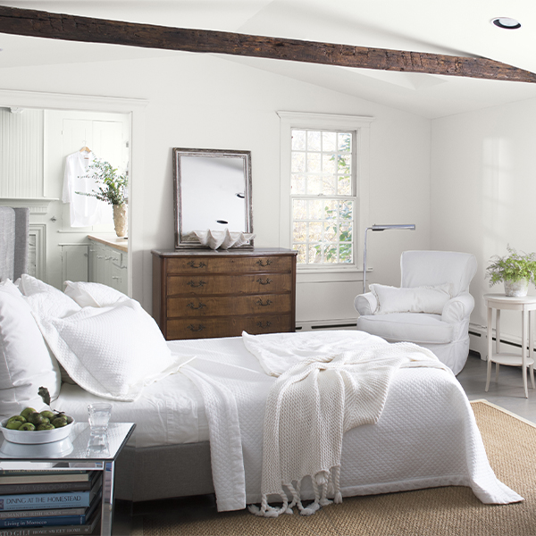 A cozy, white-painted bedroom with white bedding and chairs, a bay window with white cushions, an antique dresser, and a vaulted white ceiling with a dark brown wooden beam, looking into a white bathroom.