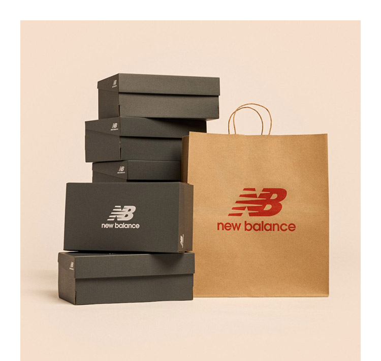 Black shoe boxes with a white New Balance logo stacked next to a brown paper shopping bag with a red New Balance logo. 