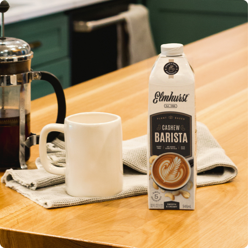 Elmhurst Cashew Barista Edition plant-based milk carton on a kitchen counter next to a French press and coffee mug.