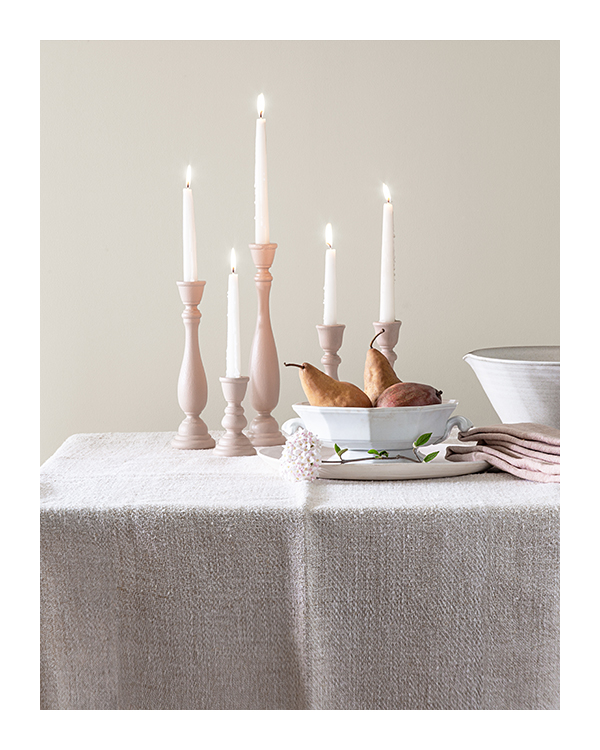 Lit tapered candles, 2 white porcelain bowls and off-white cloth napkins sit on a table against a white-painted wall.