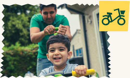 A smiling young child riding a bike with their dad behind them
