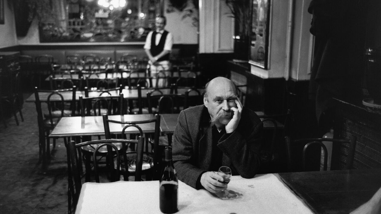 A man has a solitary glass of red wine in the Brasserie de l'Ile St. Louis. A waiter looks on in the background. 