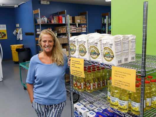 Jodi, standing in a well-stocked food pantry. Shelves display rice and cooking oil. Jodi is smiling at the camera.