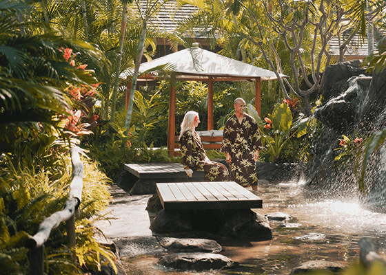 A couple in matching robes standing in a lush oasis with vegetation, waterfalls, and seating areas.