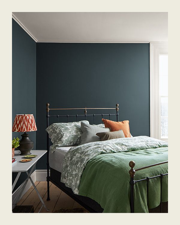 A dark teal-painted bedroom with a metal bedframe topped with a mattess, green linens, and a trio of throw pillows.