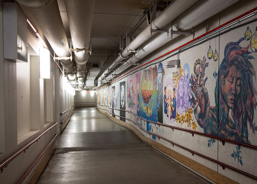 Underground tunnel with pipes at ceiling, bright lights, siderails, and a mural-decorated wall on right.