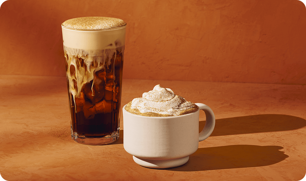 A Pumpkin Cream Cold Brew in a glass sits next to a Pumpkin Spice Latte in a short white mug on an orange background.