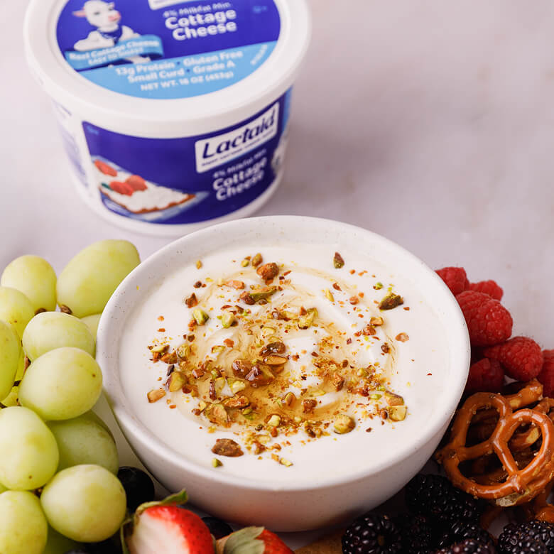 Whipped cottage cheese dip on a plate with a medley of fruit and pretzels with tub of cottage cheese nearby.