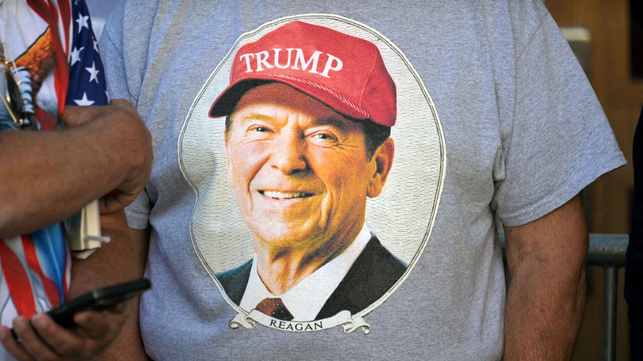 A man wears a t-shirt with a picture of former US president Ronald Reagan wearing a Trump hat.